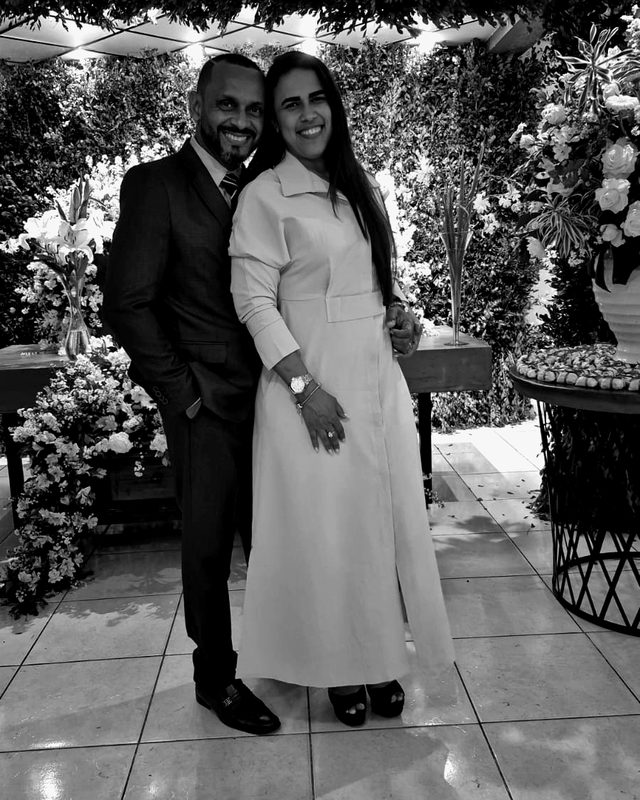 Pastor Evandro e Pastora Simone, abraçados, em frente a um painel de folhagens e flores brancas.
